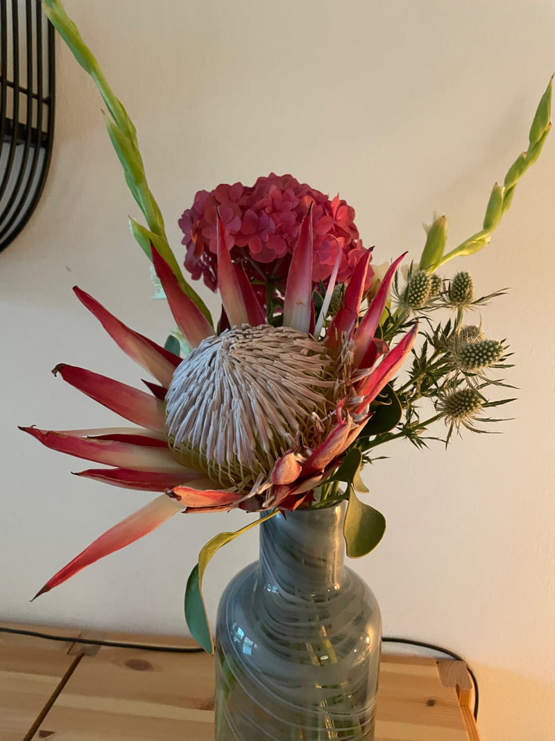 Widoczny jest duży kwiat protezy królewskiej (Protea cynaroides), będący częścią kolorowego bukietu w szklanym wazonie. Charakterystyczne, spiczaste podkwiat...